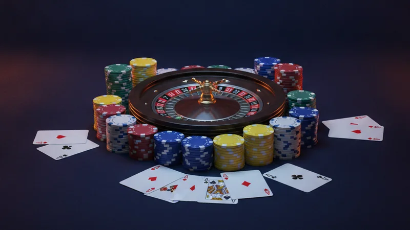 Online casino roulette wheel, playing cards, and chips on a dark navy felt table with blue and orange lighting.